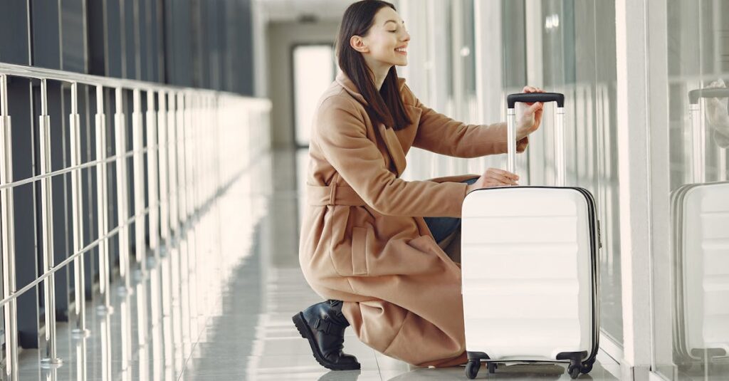 Side view full length of cheerful young female passenger in stylish beige coat with suitcase hunkering down and looking out glass wall in contemporary airport corridor